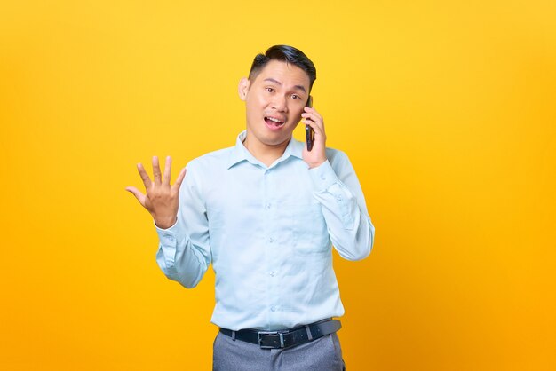 Hombre de negocios guapo joven sorprendido hablando por teléfono inteligente y mano levantada sobre fondo amarillo