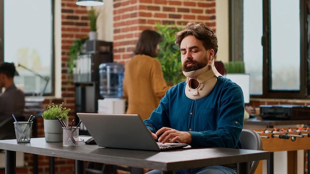 Hombre de negocios herido usando un aparato ortopédico cervical en el trabajo de oficina, trabajando en un proyecto con dolor y lesiones. Persona con tensión física y espuma de cuello tratando de curar después de un accidente, sufriendo en el trabajo.