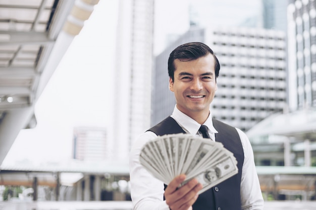 Hombre de negocios hermoso de la cara feliz que lleva a cabo billetes de dólar de EE. UU. Del dinero en el distrito financiero urbano