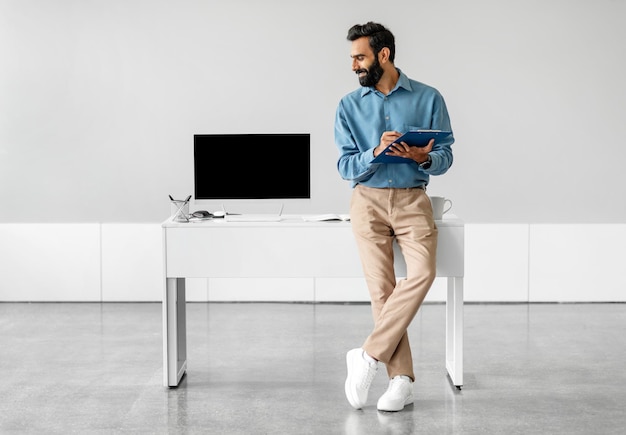 Un hombre de negocios indio confiado sentado apoyado en la mesa y mirando la computadora con una pantalla en blanco