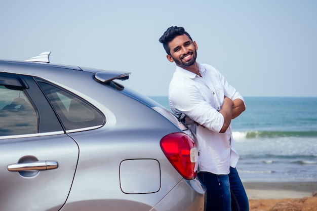 Hombre de negocios indio parado cerca del auto al aire libre en la playa del mar verano buen día un hombre con una camisa blanca y una sonrisa blanca como la nieve regocijándose comprando un auto nuevo disfrutando de unas vacaciones junto al océano