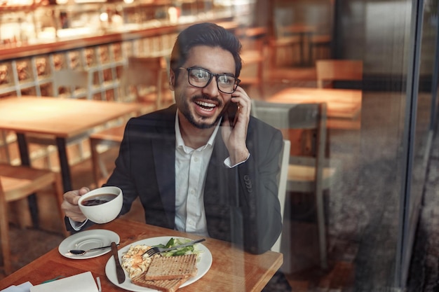 Un hombre de negocios indio sonriente está hablando por teléfono con un cliente durante la hora del almuerzo en un café acogedor