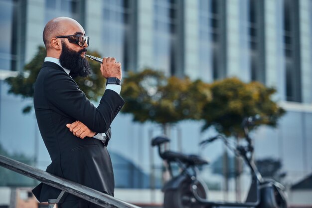 Foto un hombre de negocios inteligente y barbudo está parado cerca de su bicicleta eléctrica mientras fuma un vaporizador.