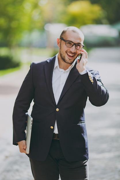 Hombre de negocios inteligente exitoso en camisa blanca, traje clásico, gafas. Hombre de pie con ordenador portátil, hablar por teléfono móvil en el parque de la ciudad al aire libre en el fondo de la naturaleza. Oficina móvil, concepto de negocio.