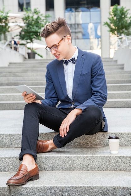 Hombre de negocios joven atractivo que se sienta con la tableta y el café en las manos en el fondo del edificio de oficinas. sosteniendo la tableta y mirando la pantalla con una gran sonrisa.