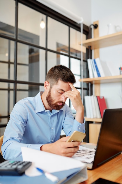 Hombre de negocios joven cansado que revisa las redes sociales en el teléfono inteligente en lugar de trabajar en la presentación del proyecto