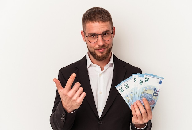 Hombre de negocios joven caucásico sosteniendo billetes aislados sobre fondo blanco apuntando con el dedo a usted como si invitara a acercarse.