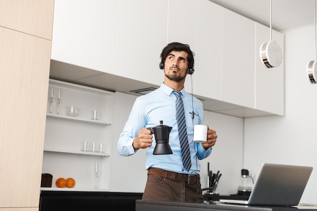 Hombre de negocios joven confidente que trabaja en casa en la cocina, usando la computadora portátil, comunicándose a través del juego de auriculares