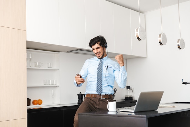 Hombre de negocios joven confidente que trabaja en casa en la cocina, usando la computadora portátil