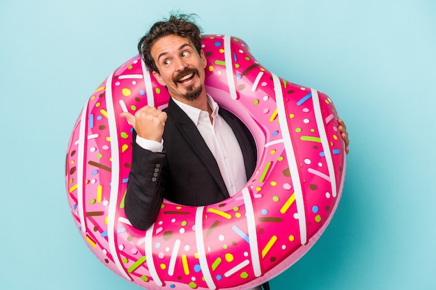 Hombre de negocios joven con donut inflable aislado en puntos de fondo azul con el dedo pulgar, riendo y despreocupado.