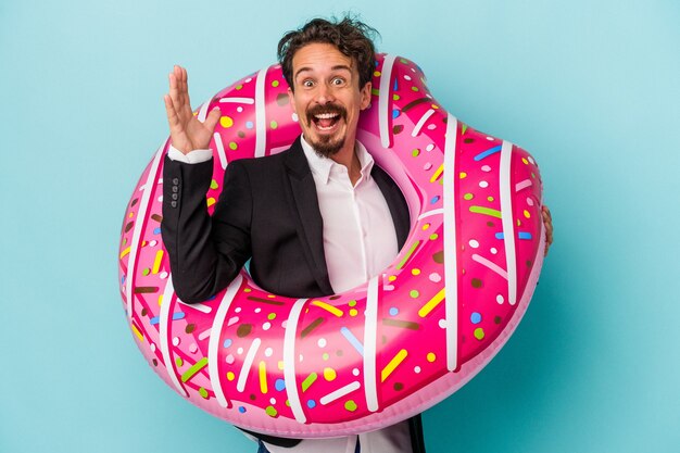 Hombre de negocios joven con donut inflable aislado sobre fondo azul recibiendo una agradable sorpresa, emocionado y levantando las manos.