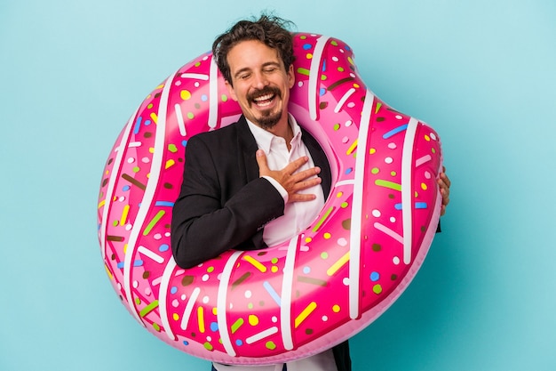 Hombre de negocios joven con donut inflable aislado sobre fondo azul se ríe a carcajadas manteniendo la mano en el pecho.