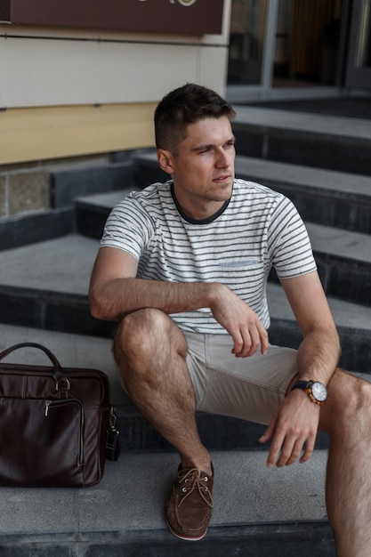 Hombre de negocios joven en una elegante camiseta de rayas de verano en pantalones cortos de moda en zapatos de gamuza con una bolsa de cuero se sienta en una escalera de piedra vintage