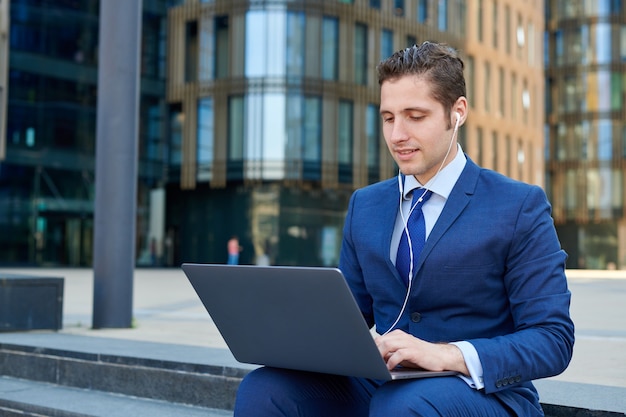 Hombre de negocios joven exitoso que trabaja en la computadora portátil moderna, usando auriculares y sentado en las escaleras afuera