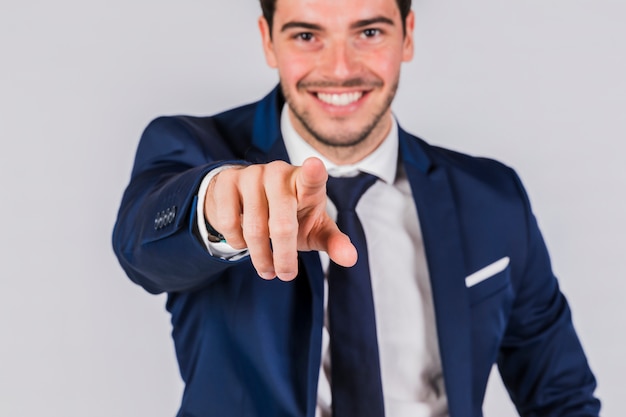 Hombre de negocios joven feliz que señala su dedo en la cámara en fondo gris
