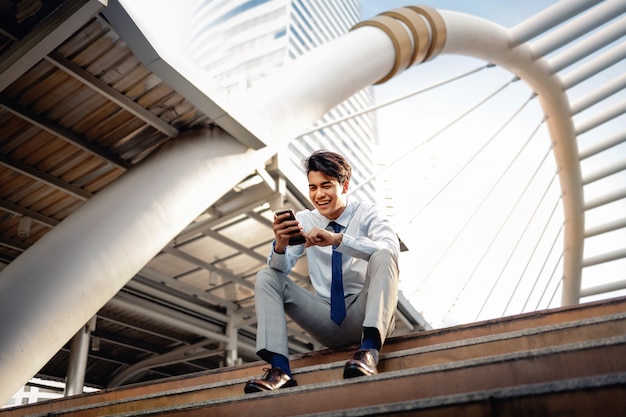 Hombre de negocios joven feliz que se sienta en escalera y que usa smartphone. Estilo de vida urbano