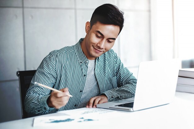 Hombre de negocios joven feliz que trabaja en la computadora portátil del ordenador en oficina. Sonriendo y escribiendo en papel