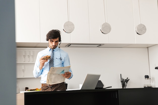 Hombre de negocios joven hermoso en la cocina comer sándwich usando la computadora portátil usando audífonos leyendo el periódico.