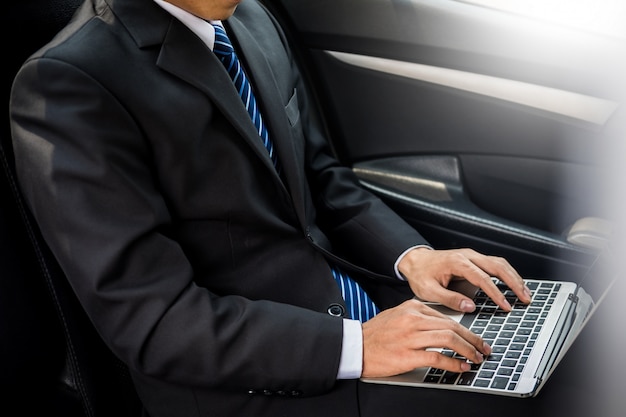 Hombre de negocios joven hermoso que usa la computadora portátil y sentándose en el asiento trasero del coche.