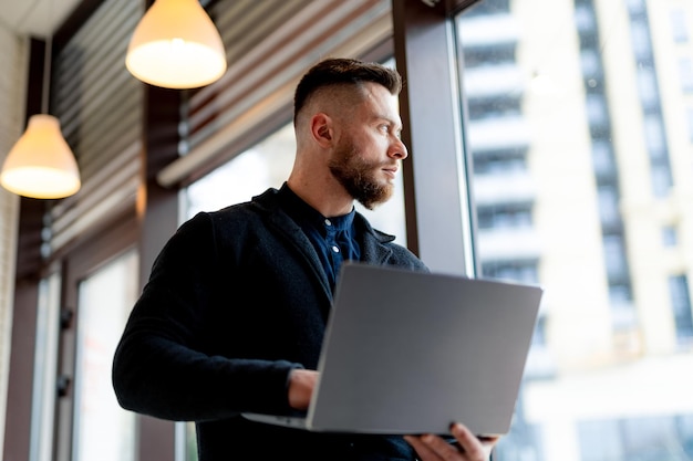 Foto hombre de negocios joven lindo trabaja en la computadora portátil de alta tecnología en un café. hombre mira la ventana con una computadora personal en las manos.