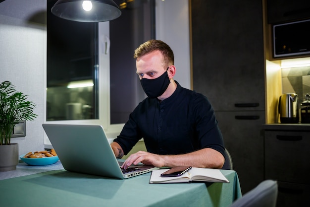 Hombre de negocios joven con una máscara protectora antivirus negra, trabajando en casa, sentado en la mesa de la cocina con una computadora portátil. Trabajar desde casa en cuarentena