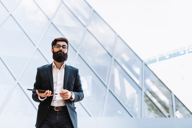 Foto hombre de negocios joven moderno que se coloca delante del edificio corporativo que sostiene la tableta digital