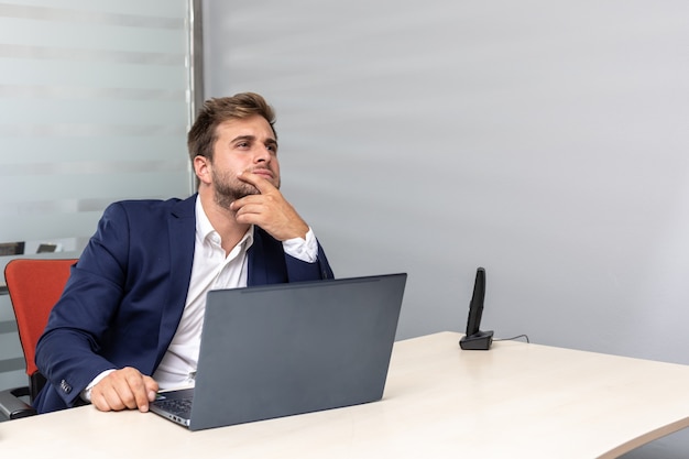 Hombre de negocios joven pensativo en la oficina sentado en el escritorio de trabajo frente a la computadora portátil.