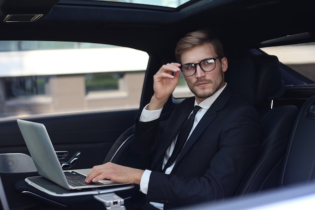 Hombre de negocios joven pensativo que mantiene la mano en los vidrios mientras está sentado en el auto lux y usa su computadora portátil.