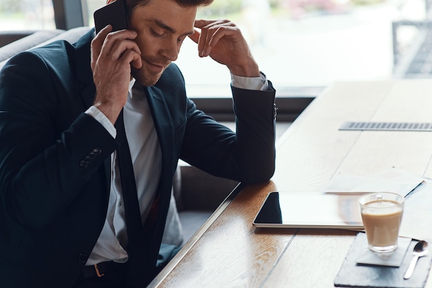 Foto hombre de negocios joven pensativo en traje completo hablando por el teléfono inteligente mientras está sentado en el restaurante