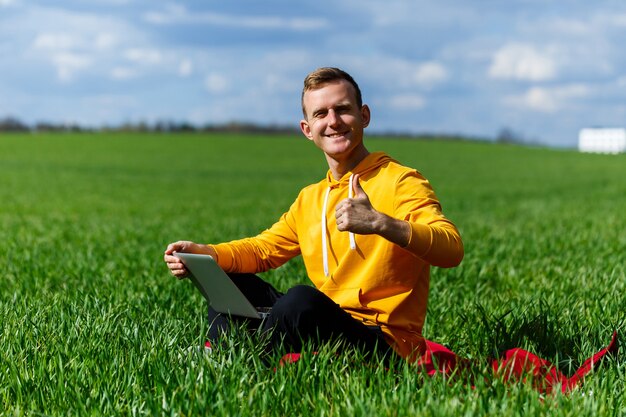 Hombre de negocios joven que se sienta en la hierba verde y que usa la computadora portátil. Hombre guapo que trabaja con la computadora en el parque en un día soleado de verano. Viaje por la naturaleza al aire libre y relajación. Concepto de trabajo autónomo