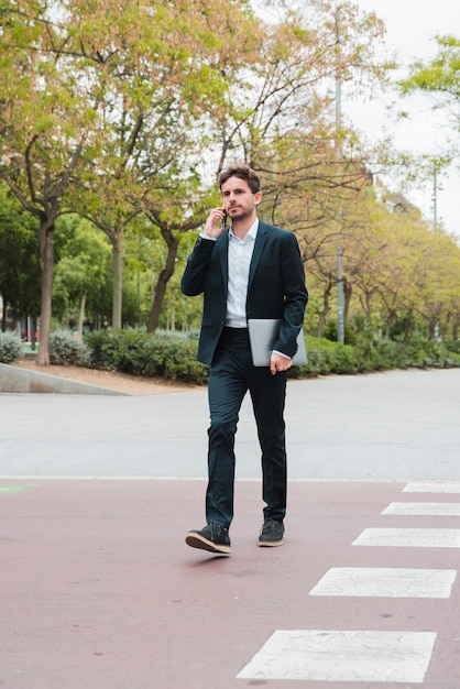 Hombre de negocios joven que sostiene el ordenador portátil en la mano que habla en el teléfono móvil que camina en el camino