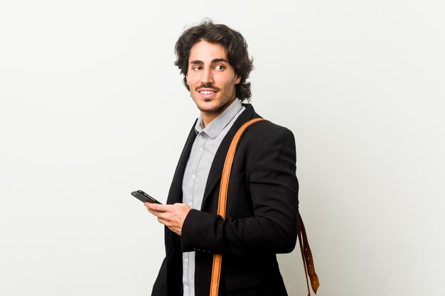 El hombre de negocios joven que sostiene un teléfono mira a un lado sonriente, alegre y agradable.