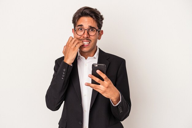 Hombre de negocios joven de raza mixta sosteniendo teléfono móvil aislado sobre fondo blanco mordiéndose las uñas, nervioso y muy ansioso.