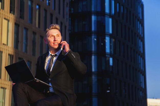 Hombre de negocios joven en ropa de desgaste de estilo, traje sentado en pasos con portátil por la noche en la puesta de sol después del trabajo. Banquero masculino relajado en la oficina de antecedentes ubicada en el distrito financiero. Copia espacio