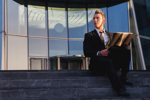 Hombre de negocios joven en ropa de desgaste de estilo, traje sentado en pasos con portátil por la noche en la puesta de sol después del trabajo