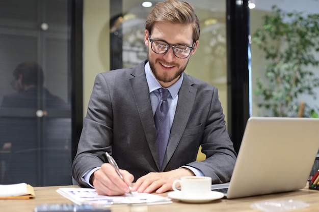 Hombre de negocios joven sonriente escribiendo algo mientras trabajaba en la oficina moderna.