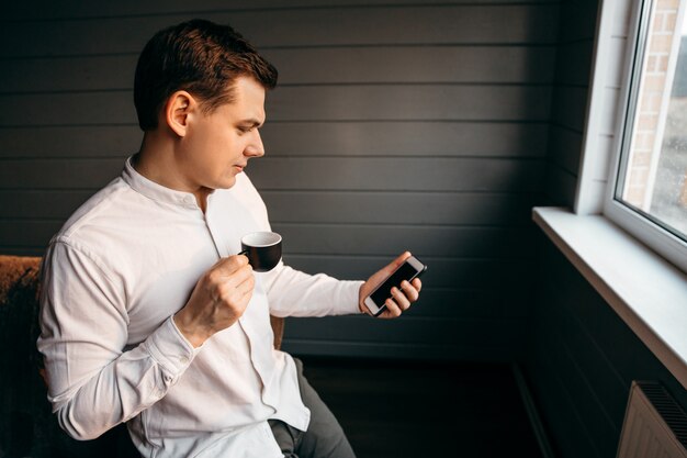 Hombre de negocios joven sonriente que usa el teléfono y bebe el café