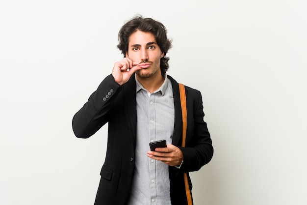 Hombre de negocios joven sosteniendo un teléfono con los dedos en los labios guardando un secreto.