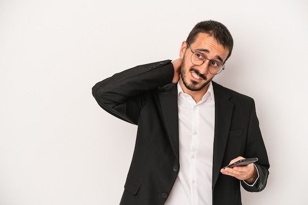 Hombre de negocios joven sosteniendo un teléfono móvil aislado sobre fondo blanco tocando la parte posterior de la cabeza, pensando y tomando una decisión.