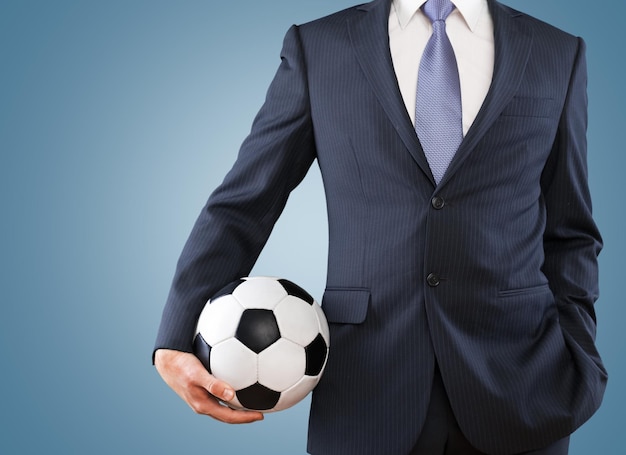 Hombre de negocios joven en traje negro con pelota de fútbol sobre fondo azul.