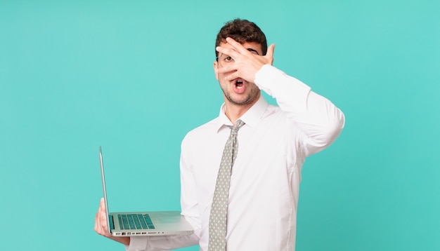 Hombre de negocios con laptop mirando conmocionado, asustado o aterrorizado, cubriendo la cara con la mano y mirando entre los dedos