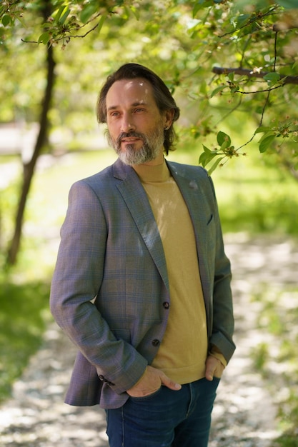 Foto un hombre de negocios maduro con barba gris con chaqueta informal y jeans pasa tiempo en el parque o en el jardín mirando pensativo a los lados de la cámara cogidos de la mano en los bolsillos