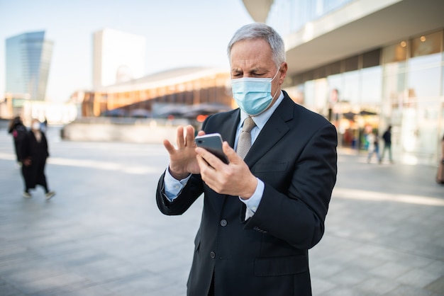 Hombre de negocios maduro enmascarado que usa su teléfono inteligente al aire libre, covid y concepto de coronavirus