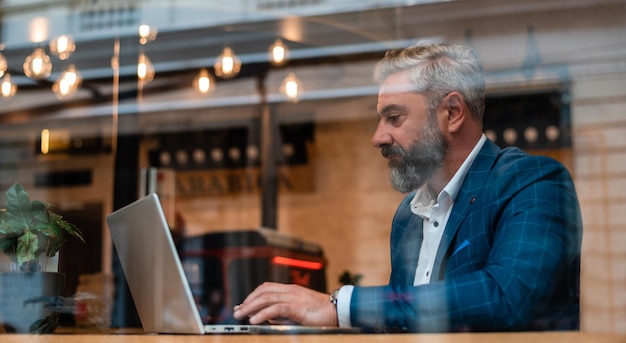 Hombre de negocios maduro sentado en su escritorio en la oficina o en la cafetería del restaurante y trabajando en una computadora portátil