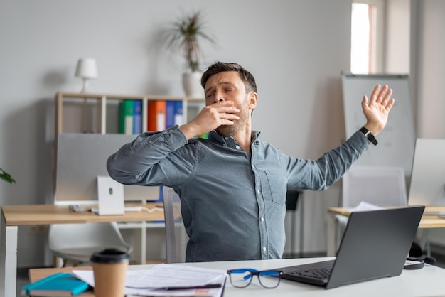 Hombre de negocios maduro soñoliento bostezando cansado de trabajar con una computadora portátil en la oficina sensación de adicto al trabajo masculino