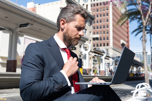 Hombre de negocios maduro en traje sentado en un banco en la ciudad al aire libre trabajando en línea en la computadora portátil, llamada de negocios.