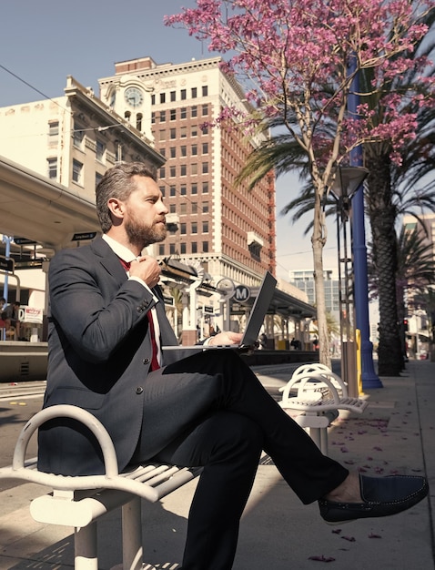 Un hombre de negocios maduro con traje se sienta en un banco en una ciudad al aire libre trabajando en línea en una entrevista de trabajo con una laptop