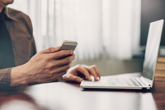 Hombre de negocios mano mensajes de texto portátil y sosteniendo el teléfono en la mesa de madera