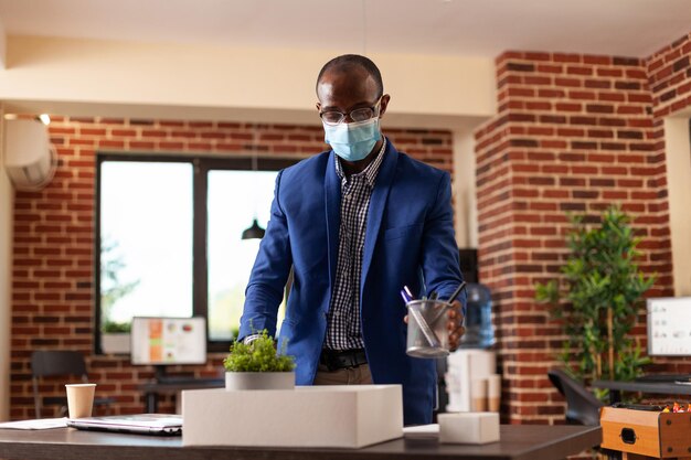 Hombre de negocios con mascarilla siendo despedido del trabajo y empacando pertenencias en caja. Empresario recogiendo cosas de oficina en el escritorio después de ser despedido y despedido de la empresa.