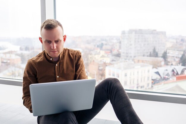 Hombre de negocios masculino exitoso joven se sienta cerca de una ventana grande con una computadora portátil. Trabajo remoto